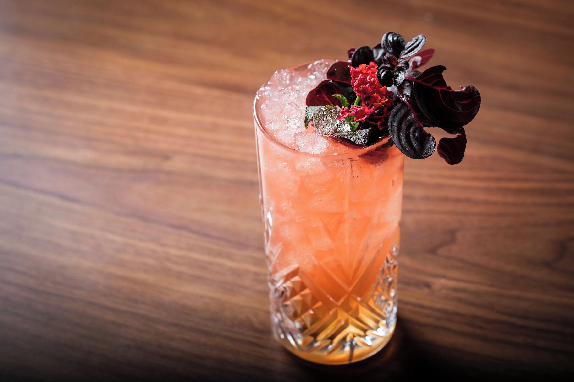 A tall glass with a pinkish drink, ice cubes, and a decorative garnish on a wooden surface.