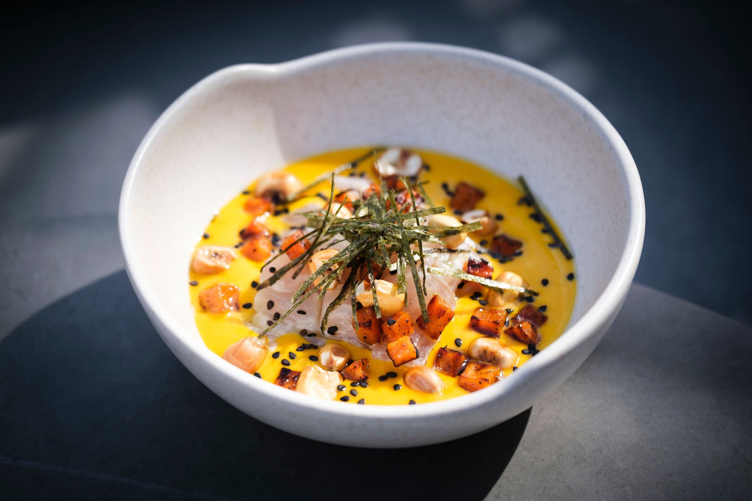A white bowl with a yellow sauce, topped with various ingredients like nuts and herbs.