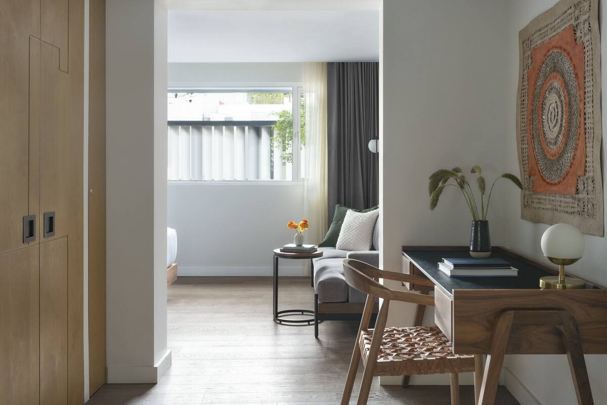 A modern, well-lit room with a wooden wardrobe, a small round table with a vase of flowers, a sofa with a green cushion, and a desk with a lamp and books.