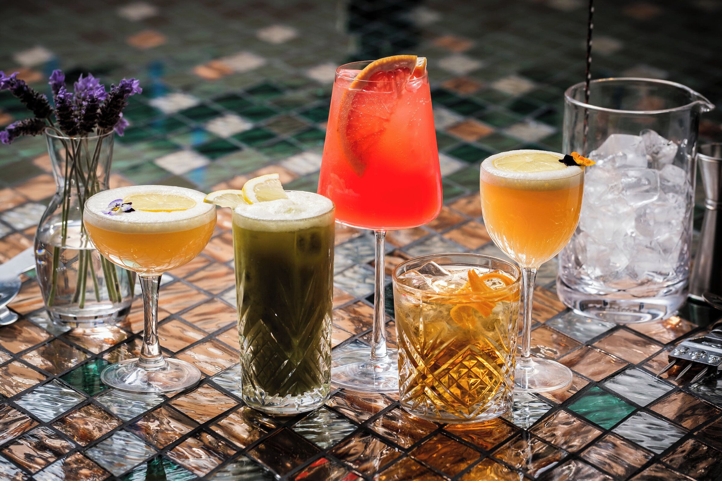 Une variété de boissons est disposée sur une table carrelée, accompagnée d’une carafe en verre et d’un vase rempli de fleurs.