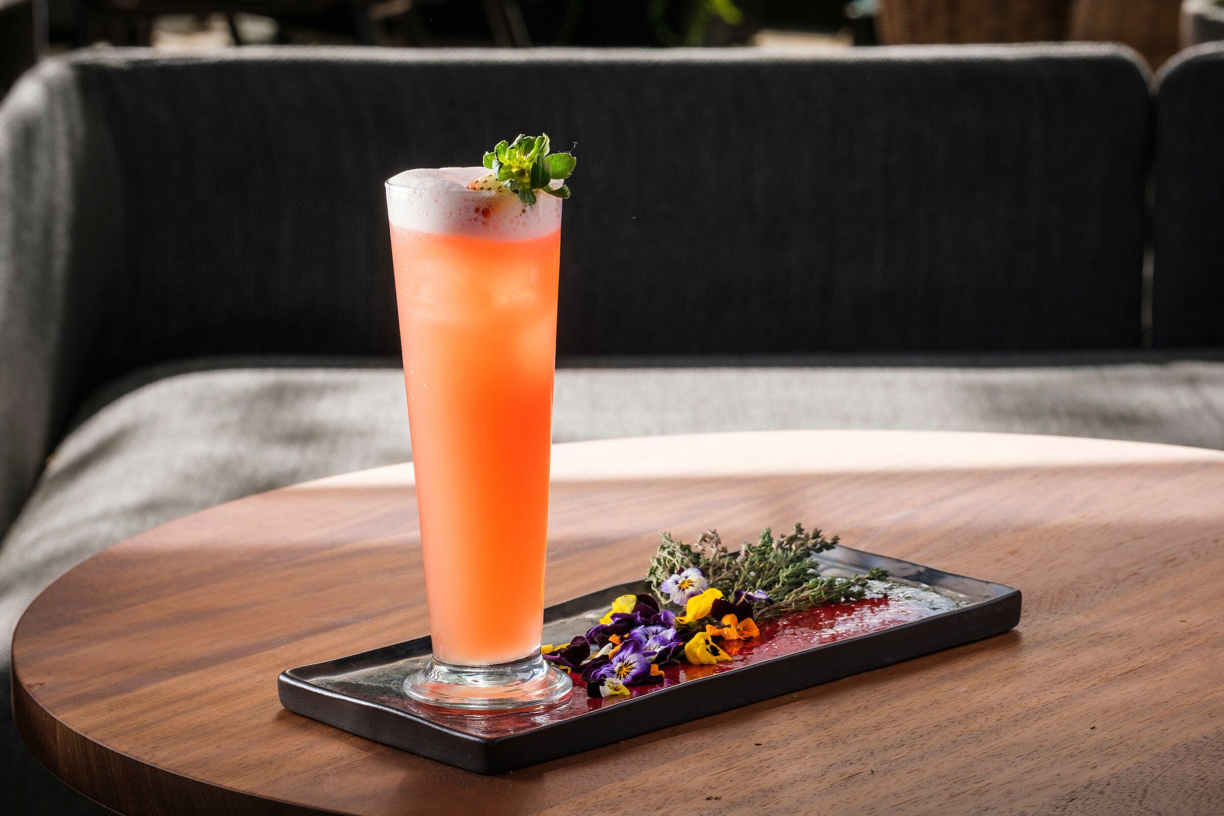 A tall glass of a pink beverage on a wooden tray with a garnish, placed on a round wooden table.