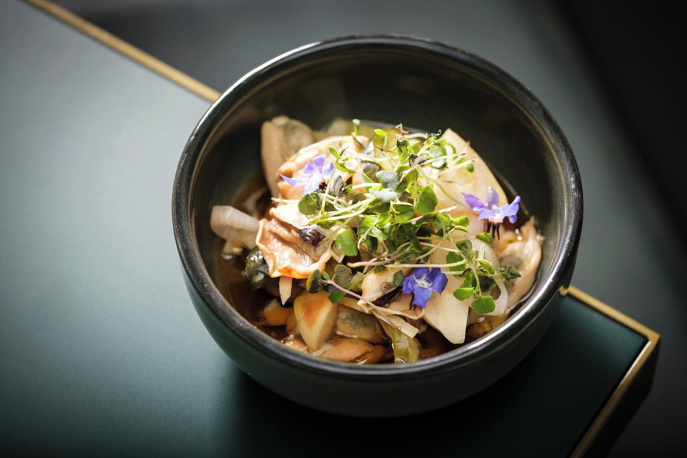 A bowl of soup with vegetables and herbs on a dark surface.