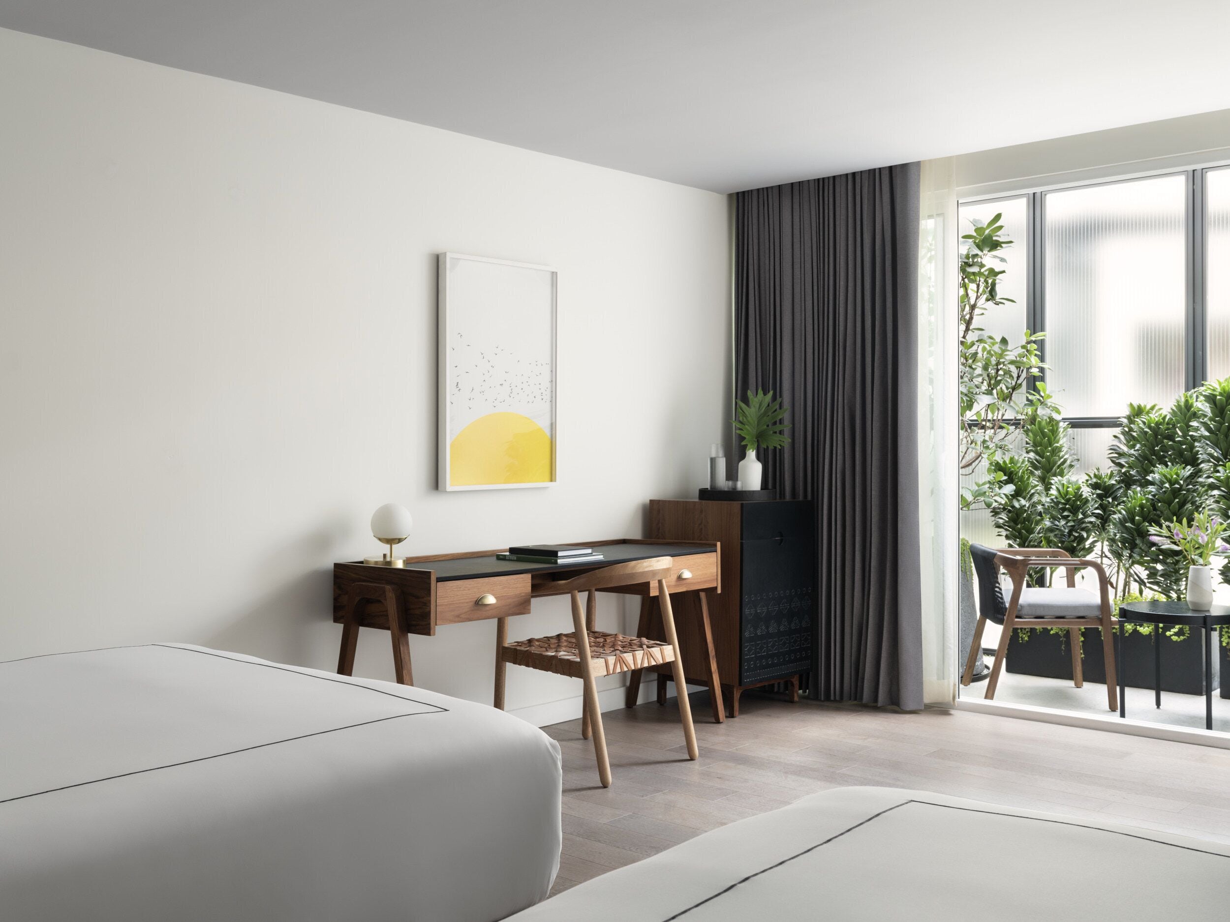 A modern bedroom with a bed, a desk, and a chair. There is a window with curtains, a plant, and a framed picture on the wall.