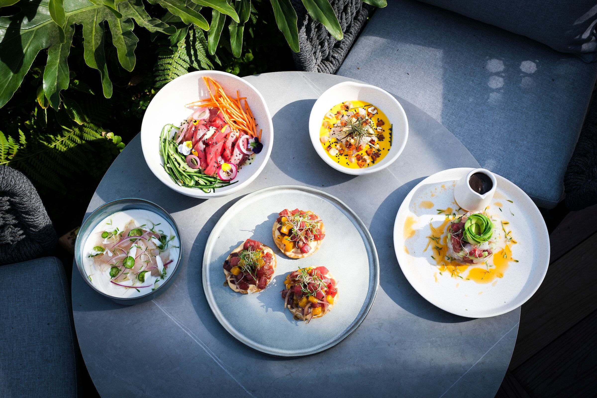 Una mesa con diversos platos, incluyendo mariscos y ensaladas, en un entorno al aire libre con vegetación de fondo.