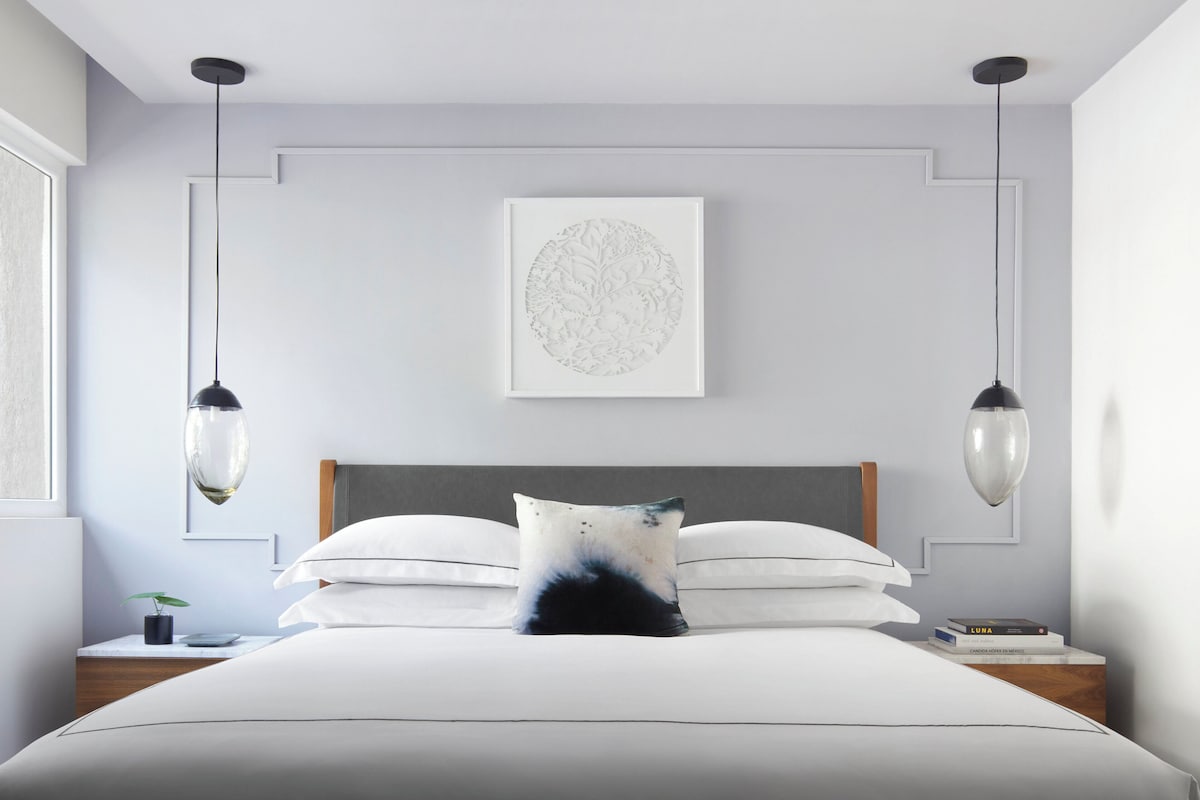 A modern bedroom with a large bed, two bedside tables, a framed artwork on the wall, and pendant lights.