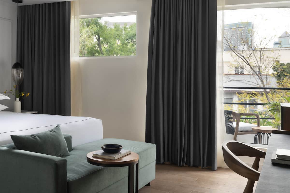 A modern hotel room with a bed, a green sofa, a small table with a bowl and book, and a view of trees and buildings through large windows.