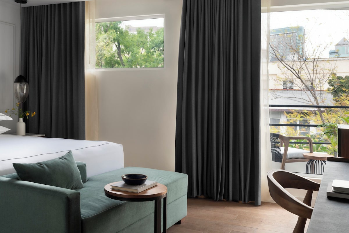 A modern hotel room with a bed, a green sofa, a small table with a bowl and book, and a view of trees and buildings through large windows.