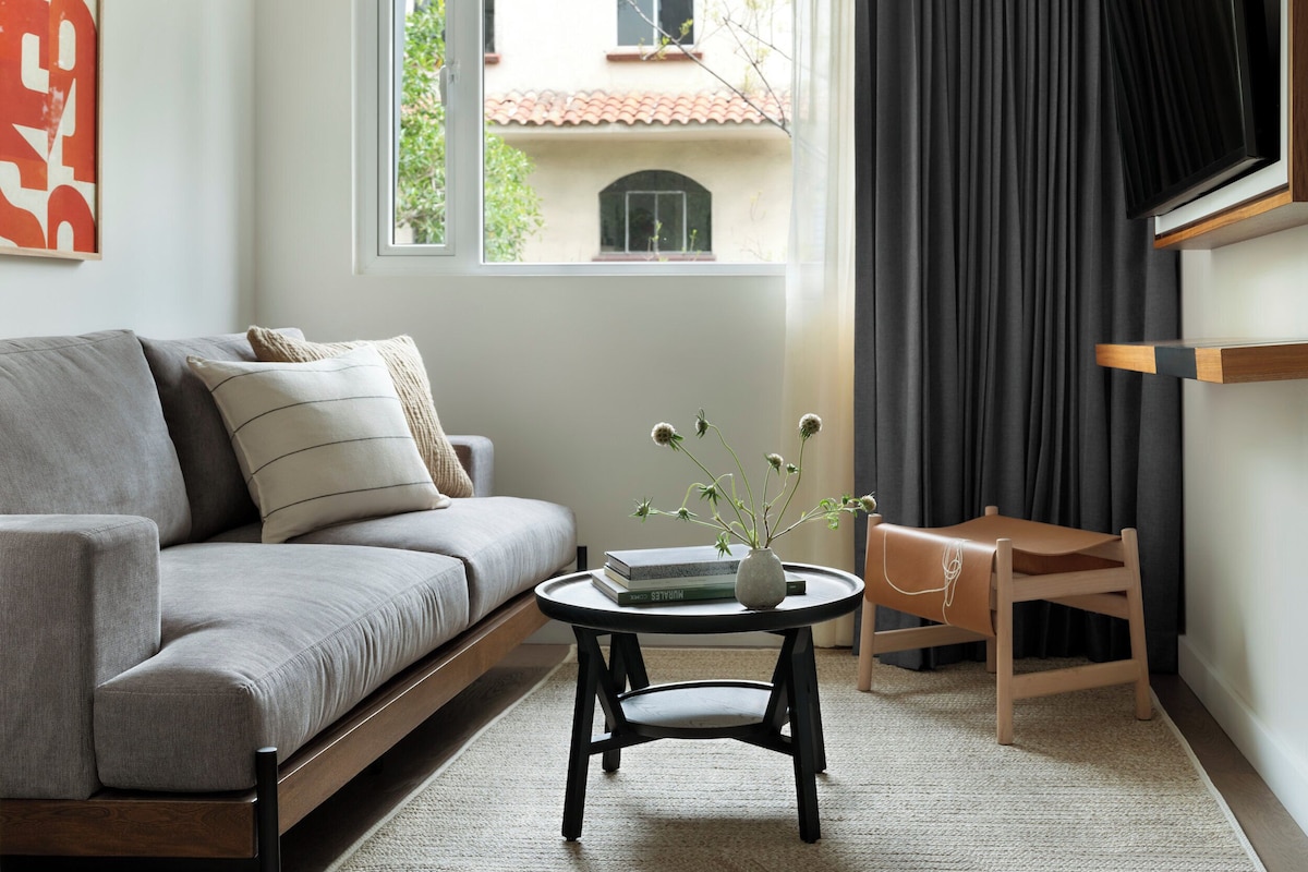 A modern living room with a grey sofa, a small round table with a vase, and a view of a building through a window.