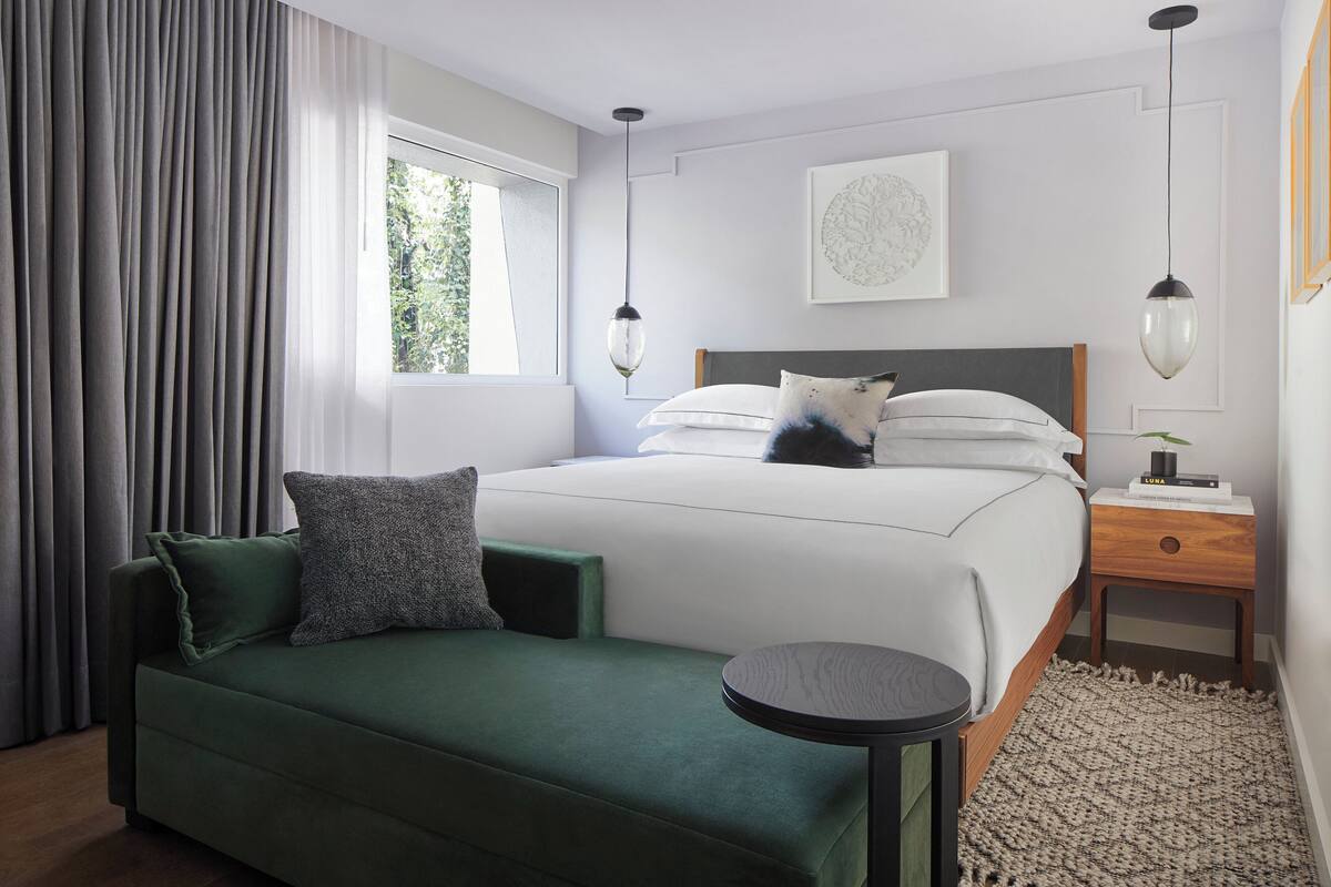 A modern bedroom with a green sofa, a round table, a bed with white bedding, and a nightstand with a lamp.