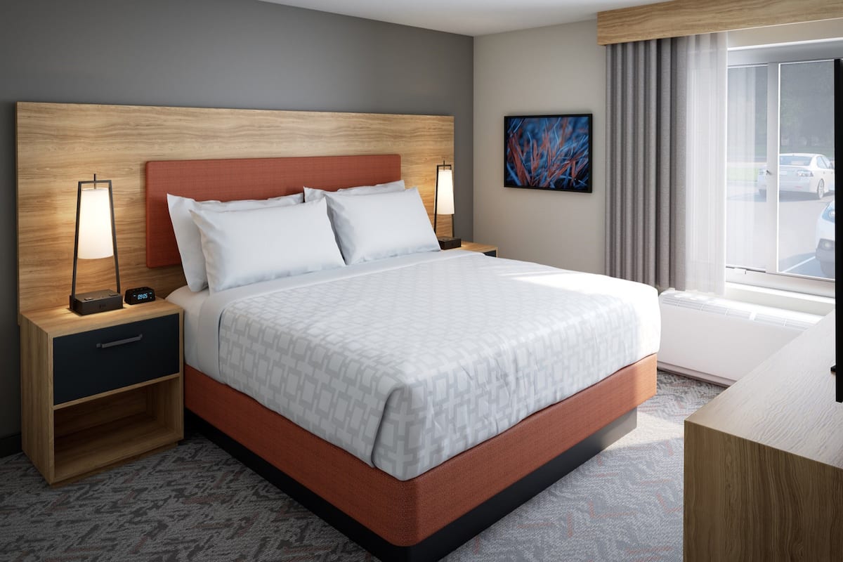 Een moderne hotelkamer met een groot bed, twee nachtkastjes met lampen, een klein plankje en een groot raam met uitzicht op geparkeerde auto's.