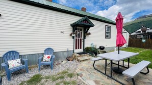 A white building with a green roof, a door with a window, and a mailbox. There is a blue chair with a floral cushion, a stone bench, and a picnic table with a pink umbrella. - The Shotgun House. (Silverton)