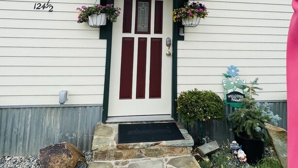 A white building with a red door and a small porch. There are flower baskets on either side of the door and a welcome sign above it. - The Shotgun House. (Silverton)