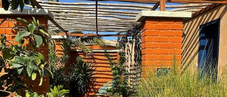 Un étroit sentier bordé de murs en briques et de plantes en pot, qui mène à une zone abritée surmontée d’une structure en bois.