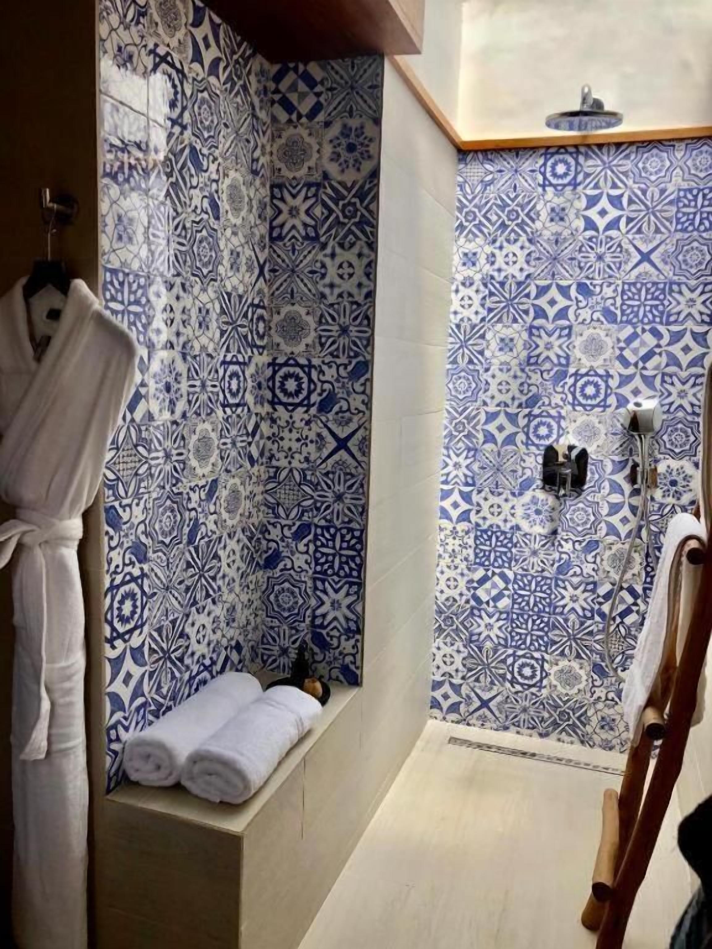 Un baño con baldosas estampadas, un banco con toallas y una silla de ducha de madera.