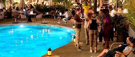 Un espace extérieur avec une piscine, des chaises longues et des tables, agrémenté de plantes en pot et illuminé de guirlandes lumineuses.