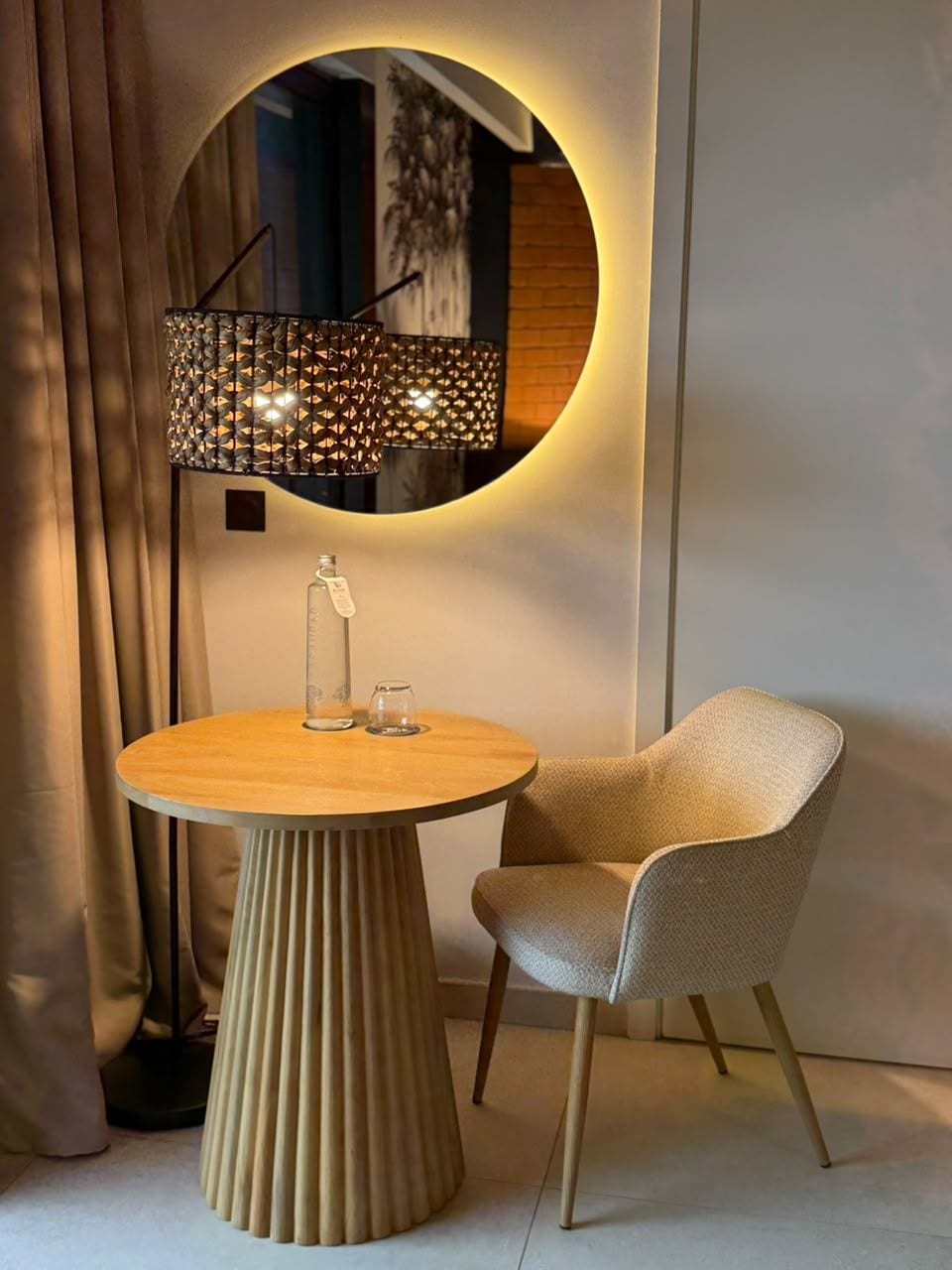 Une table ronde en bois, sur laquelle se trouvent une bouteille en verre et deux verres, un fauteuil à l’assise texturée et un luminaire mural agrémenté d’un miroir rond.