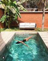 A swimming pool with a person swimming, a brick wall, a white sofa, and banana plants.