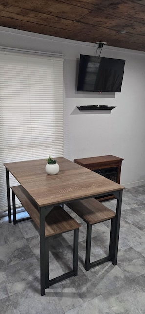 Ein moderner Essbereich mit Holztisch und Bänken, einer kleinen Topfpflanze, einem an der Wand montierten Fernseher und einem darunterliegenden Regal.