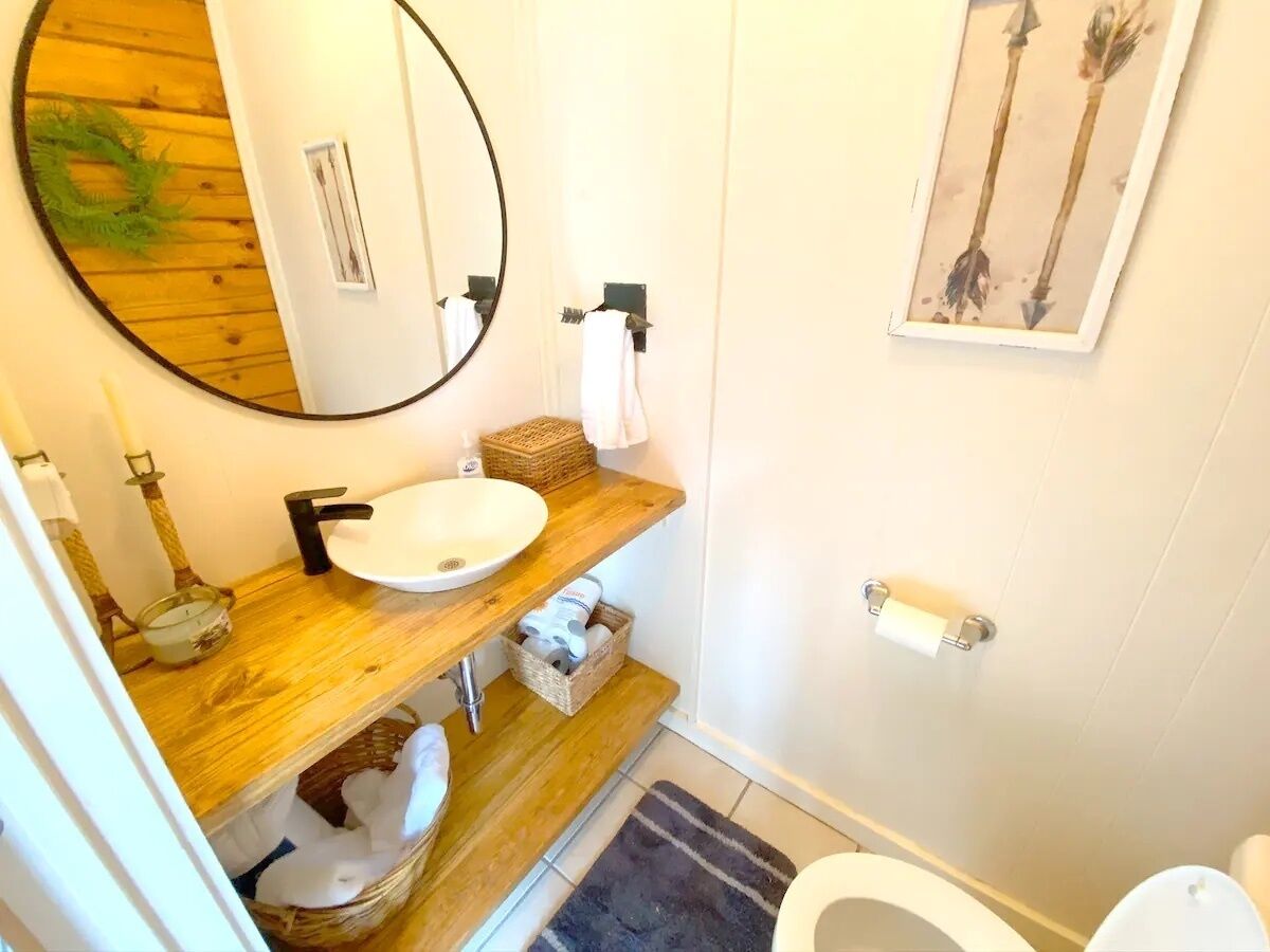 A bathroom with a wooden vanity, a round mirror, a white sink, a toilet, and a framed artwork on the wall.