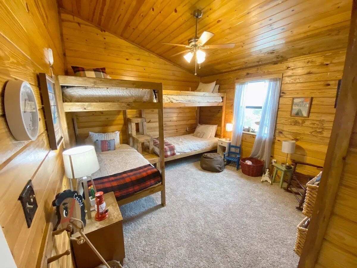 Een gezellig kamer in een chalet, met een stapelbed, een bureau met lamp, een plafondventilator en een raam met gordijnen.