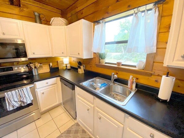A compact kitchen with white cabinets, a black countertop, a stainless steel sink, and a window with curtains.