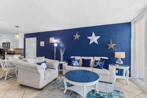 A living room with a blue accent wall, white furniture, and star decorations. - Beachfront 2BR | Private Balcony, Pool | Gulf Views (Gulf Shores)