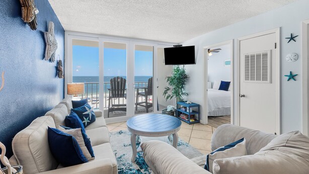 A spacious living area with a balcony overlooking the sea, a television mounted on the wall, and a bed in the adjacent room. - Beachfront 2BR | Private Balcony, Pool | Gulf Views (Gulf Shores)