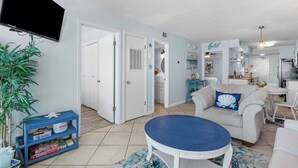 A living room with a blue round table, a sofa with a blue pillow, a bookshelf, and a TV mounted on the wall. - Beachfront 2BR | Private Balcony, Pool | Gulf Views (Gulf Shores)