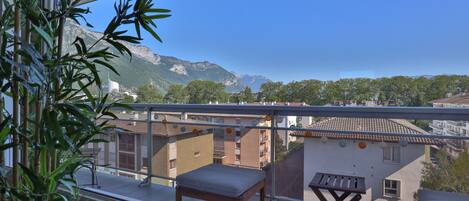 A balcony with a view of mountains and buildings, a wooden bench, and a table.
