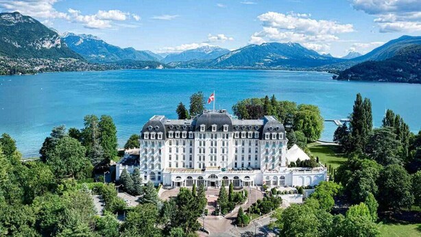 Un hotel grande e histórico con fachada simétrica, situado cerca de un lago con montañas al fondo.