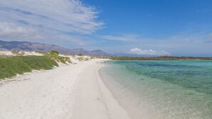 Una platja amb aigua turquesa cristal·lina, sorra blanca i vegetació verda. Al fons, es veuen muntanyes.