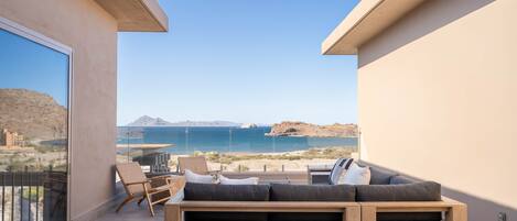 A balcony with a sofa, chairs, and a glass railing overlooking a coastal landscape.