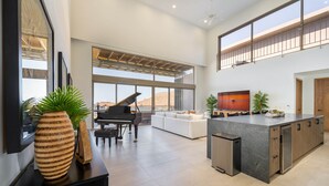 A spacious living area with a piano, a large window, a modern kitchen, and a balcony.