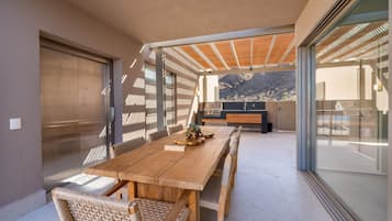 Uma área de refeições moderna ao ar livre, com mesa de madeira e cadeiras de vime, porta de vidro deslizante e vista para montanhas.