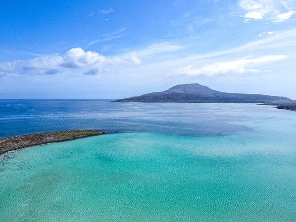 A coastal landscape with clear turquoise waters, a distant mountain, and a rocky shoreline.