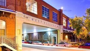 Edifício de tijolos com a inscrição "WEST VILLAGE" na fachada, apresentando uma área de estacionamento coberta e carros estacionados por baixo.