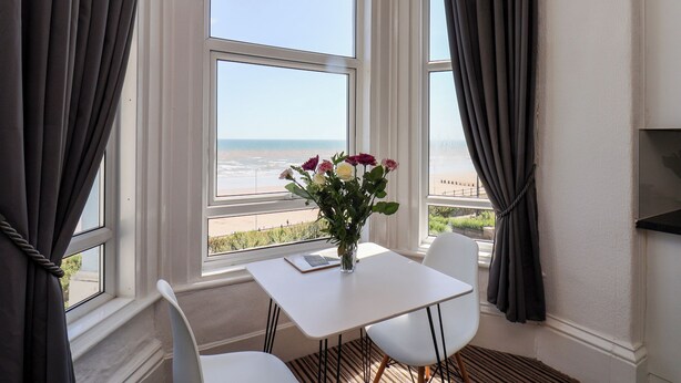 A room with a view of the beach, a white table, two chairs, and a vase with flowers. - 6 Beach View @ Beaconsfield House (Bridlington)
