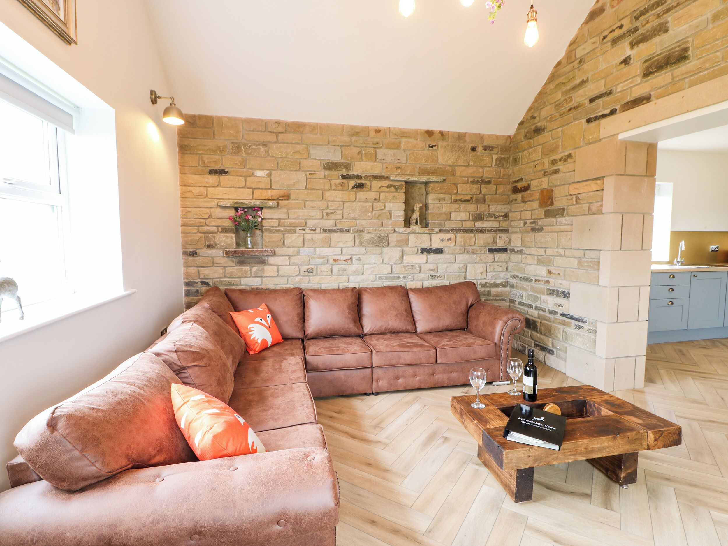 A living room with a brown leather sectional sofa, a wooden coffee table with wine glasses, and a stone brick wall.