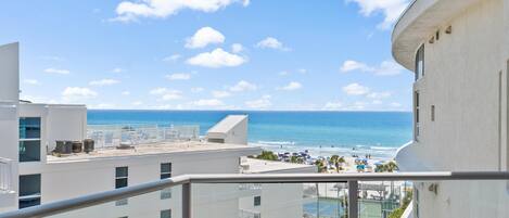 A balcony with a view of the beach, a tennis court, and a pool area.