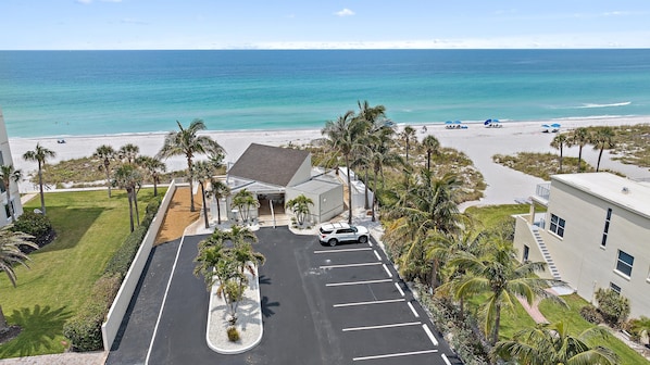 A parking lot with a white car, surrounded by palm trees and a building with a grey roof.
