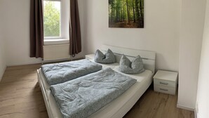 Quarto com cama, duas almofadas, mesa de cabeceira, janela com cortinas e arte na parede com tema de floresta.