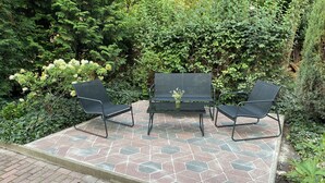 Um pátio externo com azulejos de padrão hexagonal, rodeado por vegetação exuberante e diversas plantas.