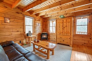 A cozy cabin interior with a wooden floor, a leather sofa, a wooden table, a fireplace, and large windows. - The Hemlock | Hocking Hills, Romantic, Hot Tub (Rockbridge)
