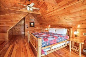 Camera da letto in una baita di legno con soffitto spiovente, un letto con trapunta colorata, una ventola a soffitto e un comodino con una lampada.