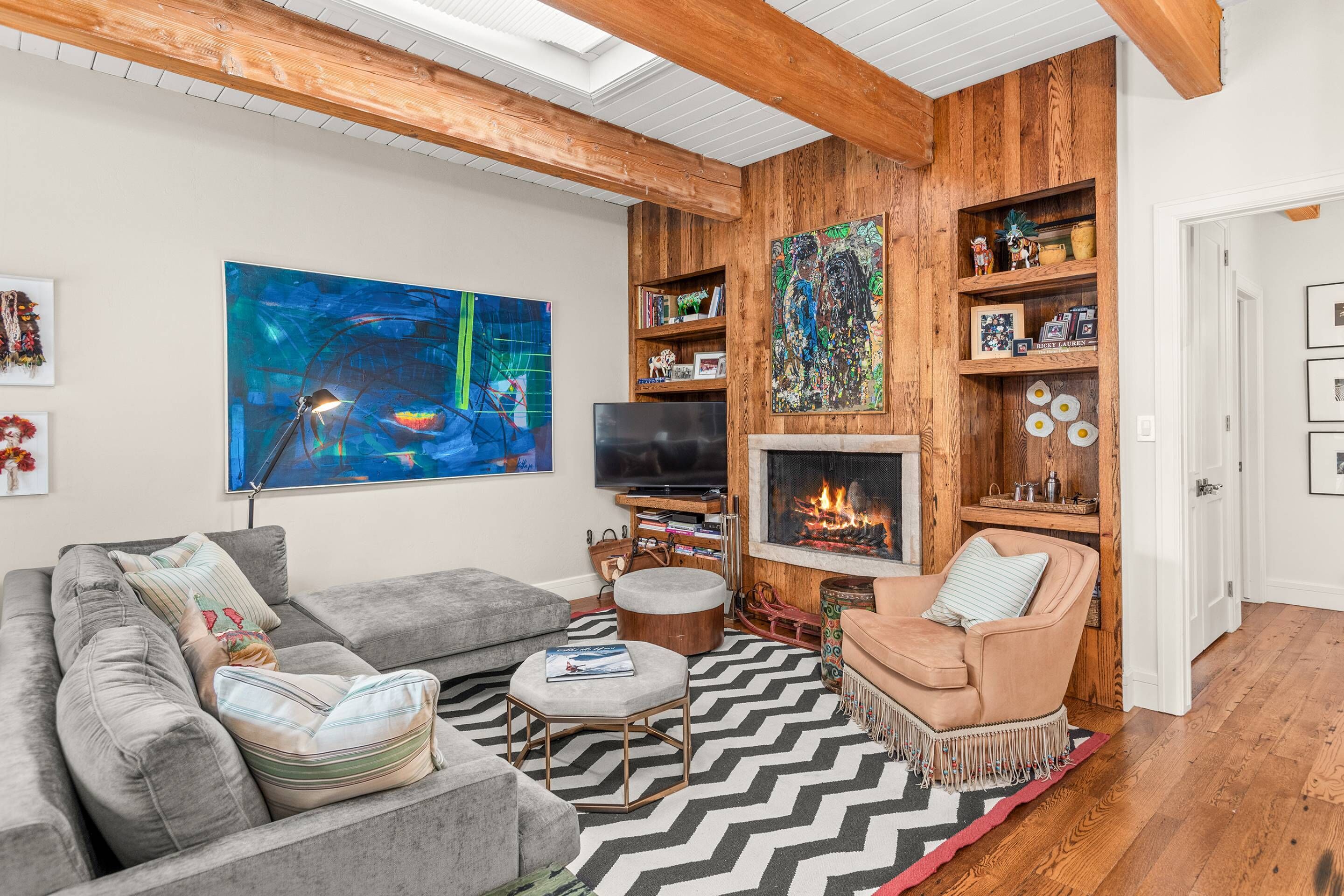 A living room with a grey sofa, a round ottoman, a beige armchair, a fireplace, wooden shelves, and a large abstract painting.