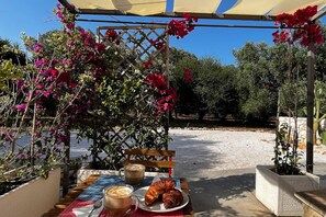 Un área de estar al aire libre con una mesa para dos, un banco de madera y una pérgola con plantas florecientes.