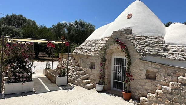 A stone building with a conical roof and a small door, surrounded by potted plants and a pergola. - Trullo Oscar (Ceglie Messapica)