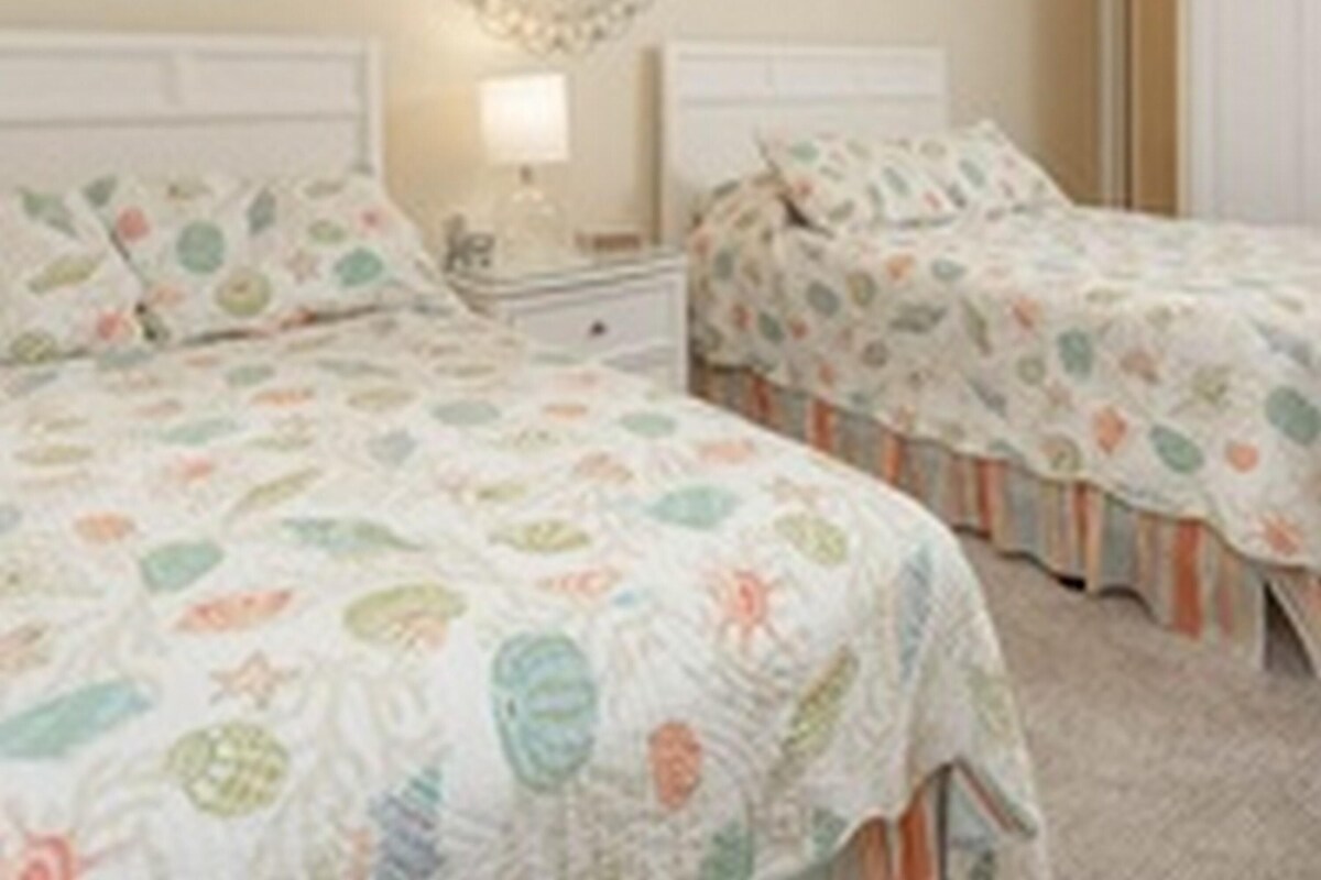 Quarto com duas camas, uma mesa de cabeceira, um objeto decorativo na parede e um ventilador de teto.