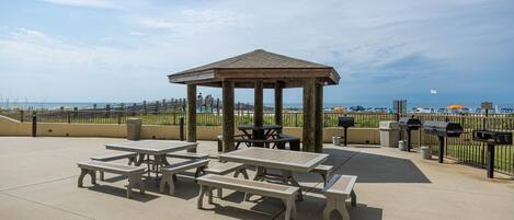 Un espace pique-nique abrité, équipé de tables et de bancs, avec un barbecue et une vue sur la plage.