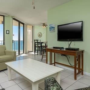 A living room with a sofa, a coffee table, a TV on a wooden stand, and a view of the sea through the glass door.